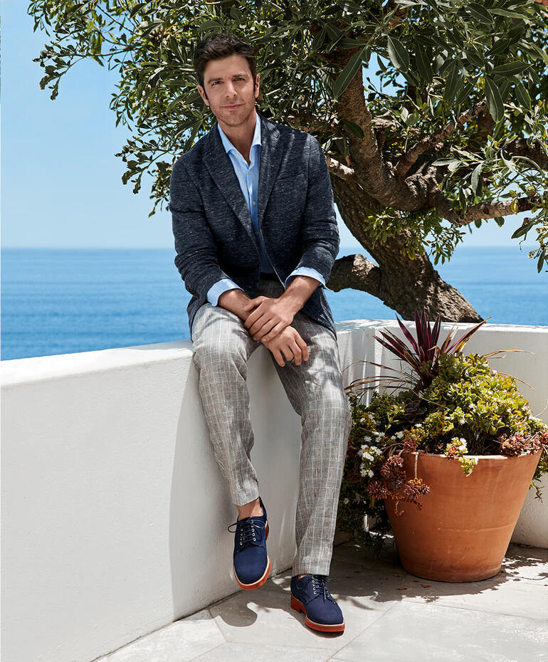Bottoli’s linnen glencheck-pantalon Man in Barutti linnen jersey blazer, zittend aan een muur bij de zee.
