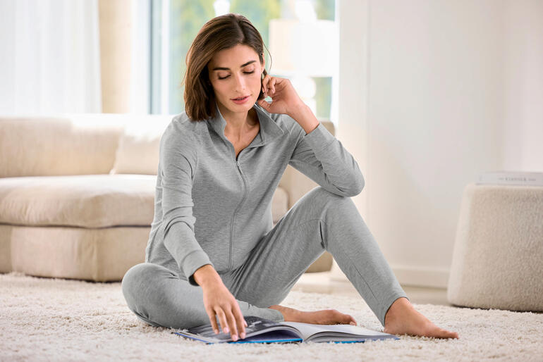 Vrouw zit in een grijs modal huispak en leest een boek.