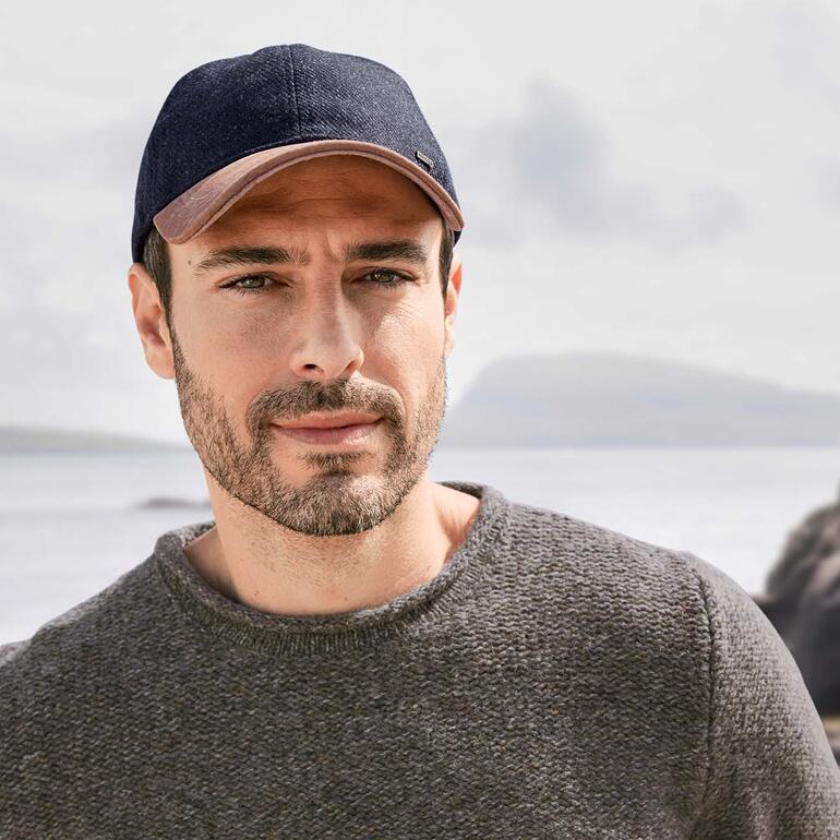 Man in een visserspullover met een donkerblauw pet, kijkend naar een waterview.