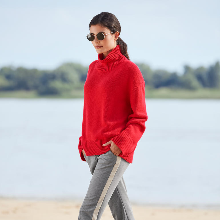 Seductive galonbroek ‘Blended Wool’ Vrouw in een rode trui met galonstrepen, elegant langs het water wandelend.