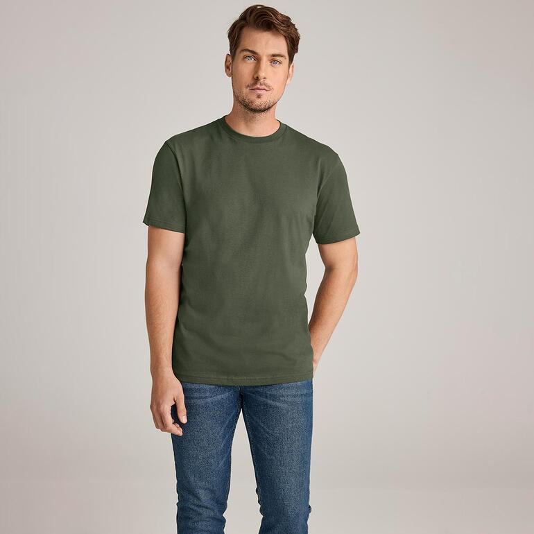 Man in olijfgroen T-shirt met jeans, staand voor een neutrale achtergrond.