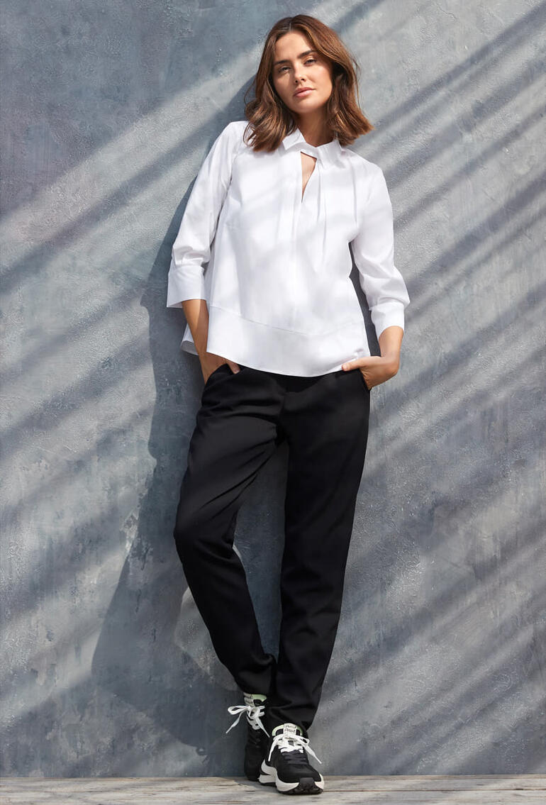 RAPHAELA BY BRAX onderhoudsarme broek Vrouw in witte blouse en zwarte broek staat tegen een grijze muur
