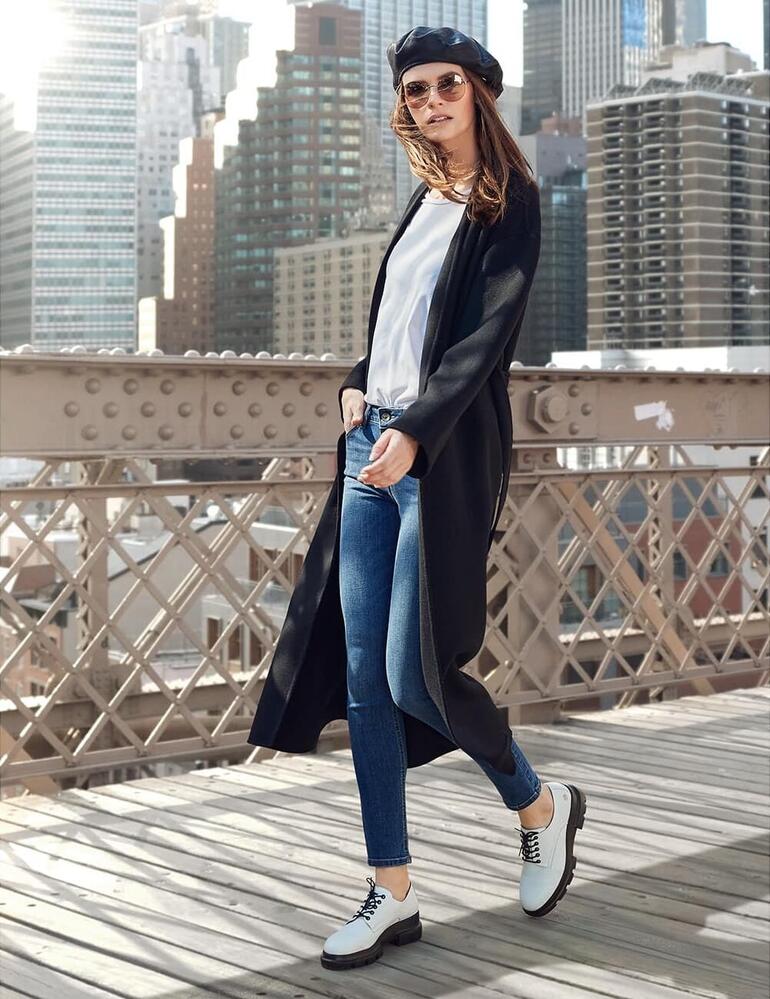 wit Model in een zwarte mantel met jeans in stadslandschap.