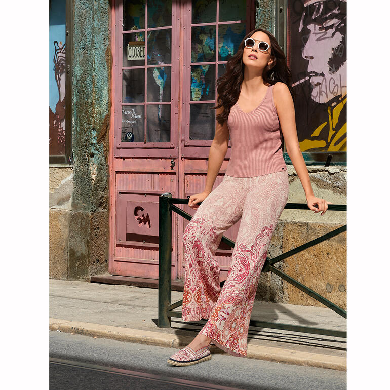 Ivko gebreide espadrilles Vrouw in roze top en kleurrijke broeken zit voor een kleurrijke muur.