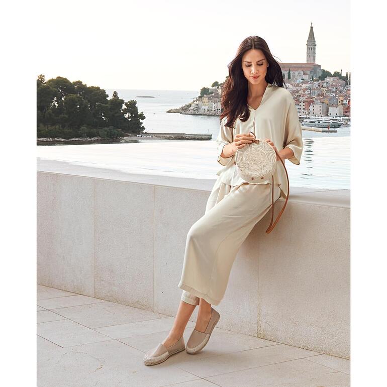beige Vrouw zit op een muur, in lichte kleding en leren espadrilles.