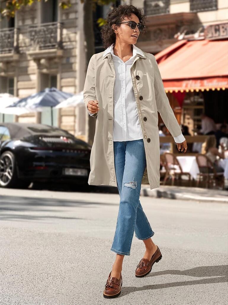 Vrouw in een beige trenchcoat en witte sneakers die door een drukke straat met cafés loopt.
