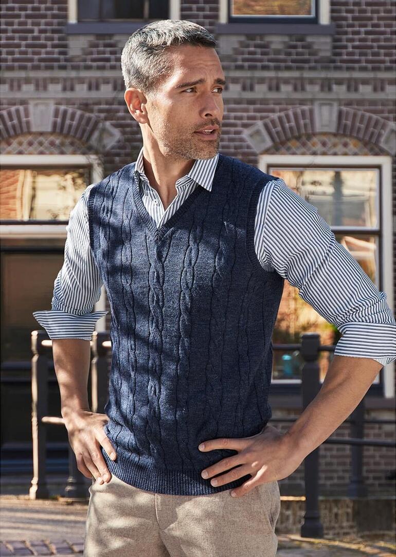 Man draagt een donkergrijze kabelvest over een shirt en staat ontspannen.