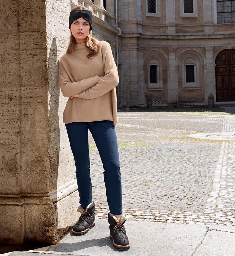 zwart Vrouw in een beige trui en blauwe jeans staat met Flufié don-sockboots op een stenen vloer.
