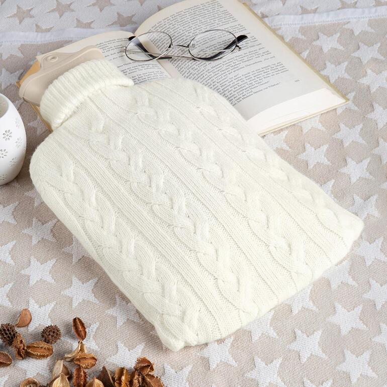 Kasjmier warmwaterkruik in crèmekleurige gebreide hoes op een tafel met een boekpagina en een bril.