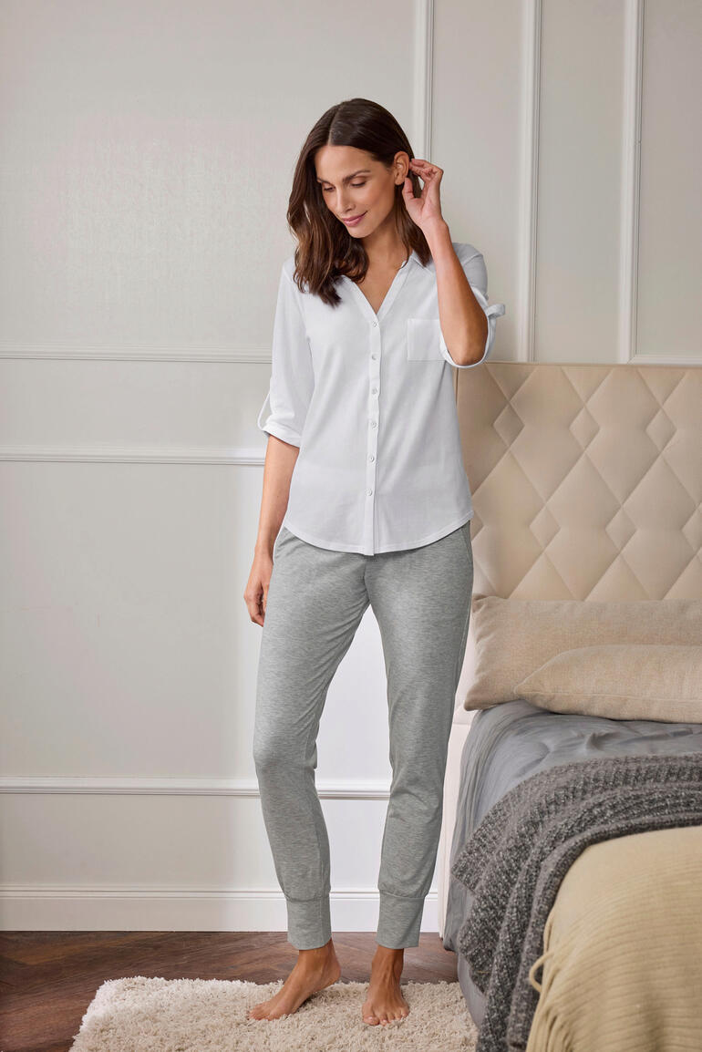 Casual pyjama Vrouw in een witte pyjama-top en grijze joggingschoenen staat naast een bed.
