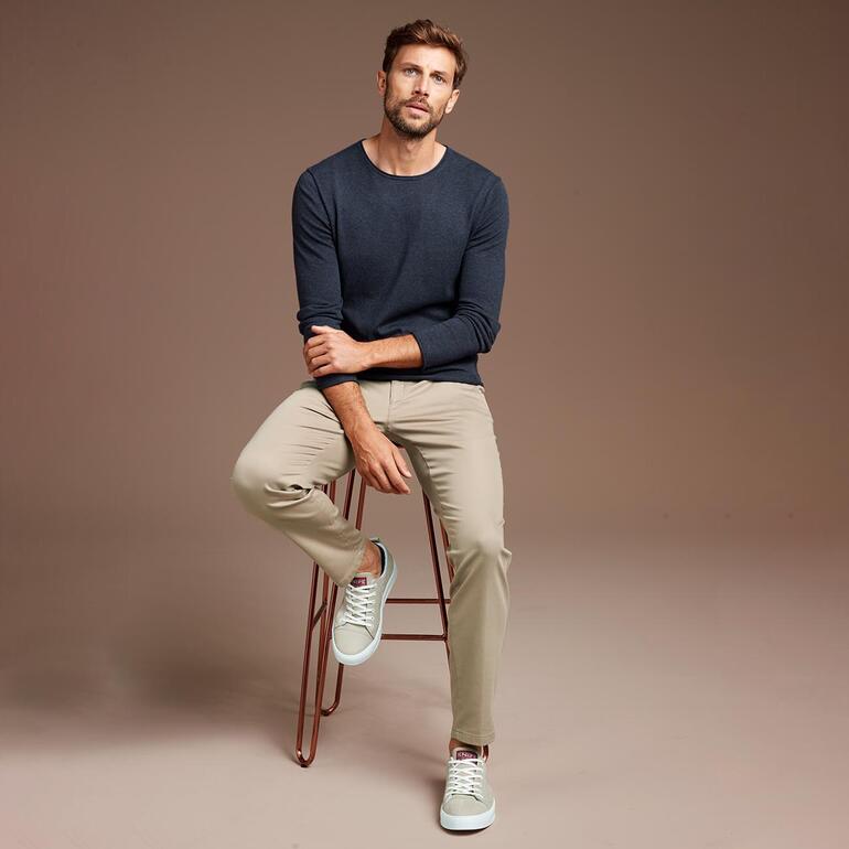 denimblauw Man zit op een barkruk, draagt een marineblauwe trui en lichte broek.