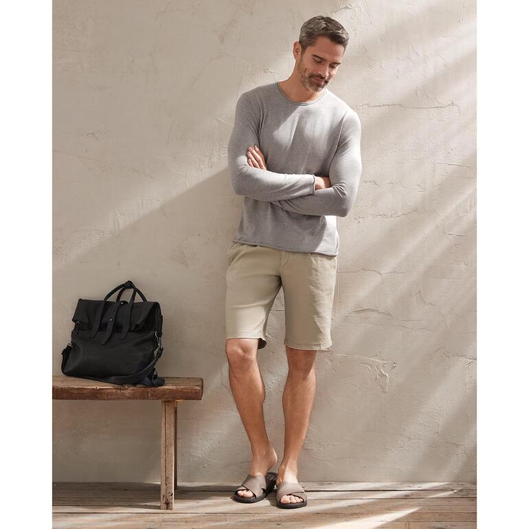 beige Man draagt lichtbeige shorts en een grijze trui, staat voor een muur.