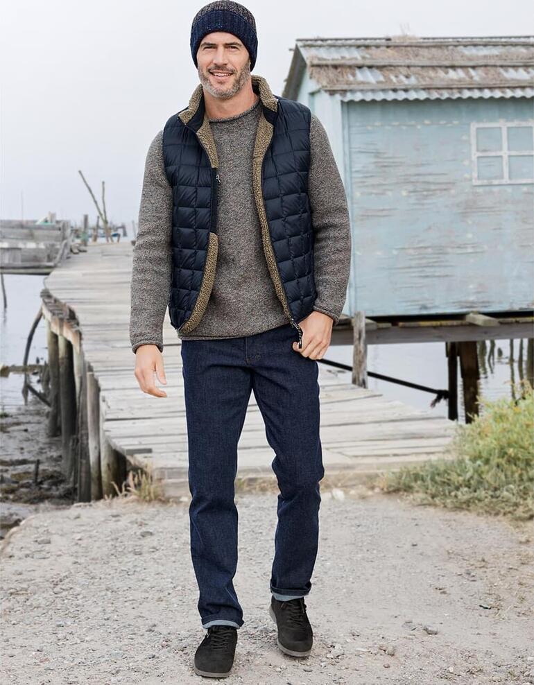 Fisherman Donegal-trui Man in een gewatteerde reversibele vest, grijze trui en muts, staat aan de haven.