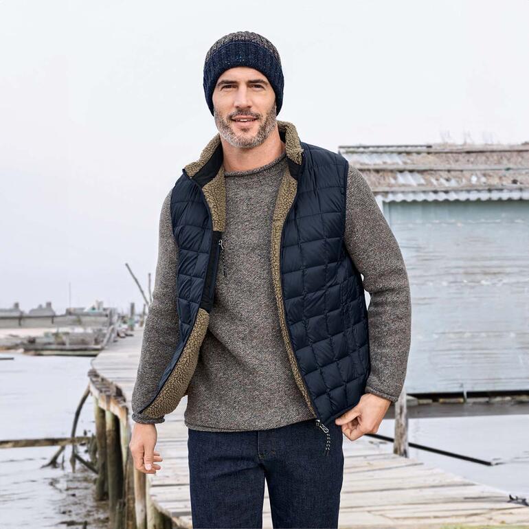 Man in gewatteerd reversibel vest poseert aan de haven met grijze trui en muts.