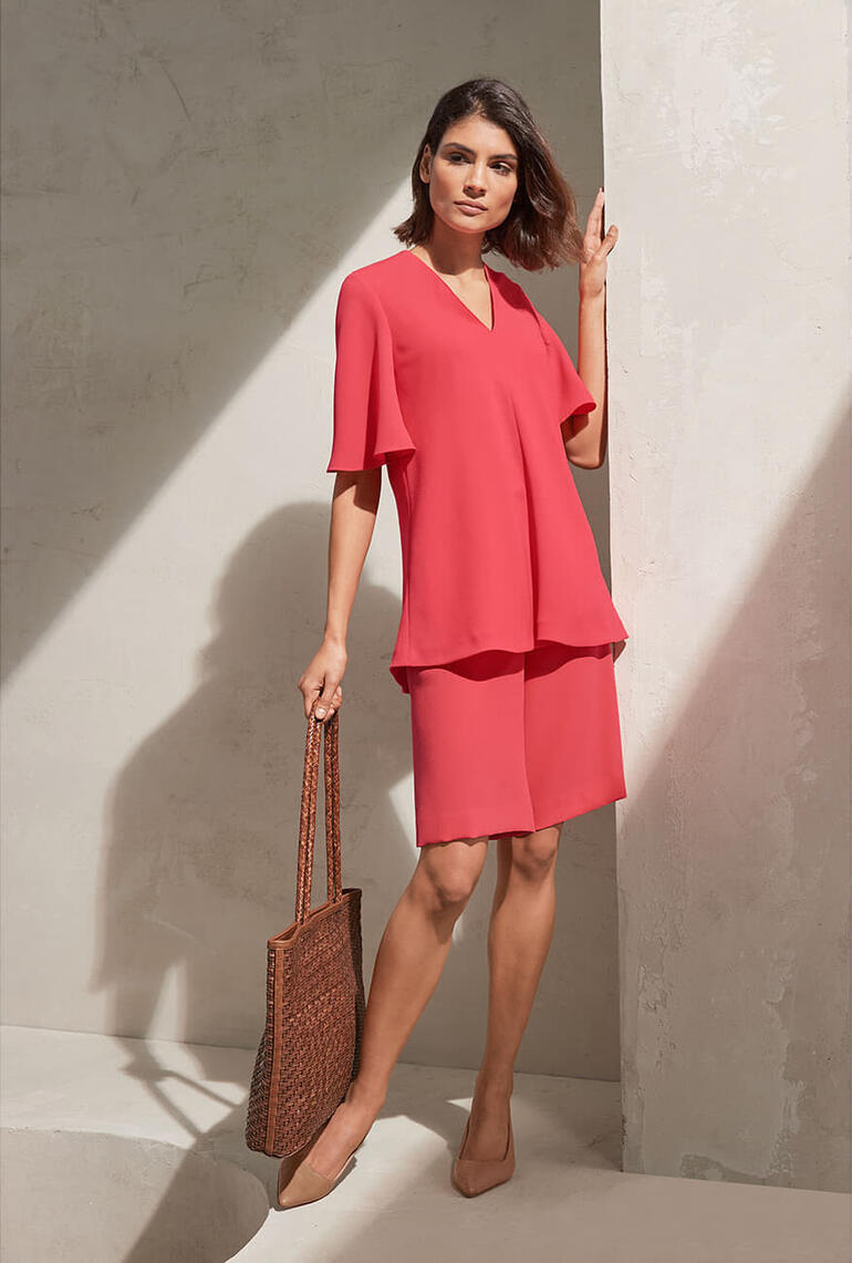 shirt met V-hals en bermudas Vrouw in een roze top en rok staat lachend voor een muur