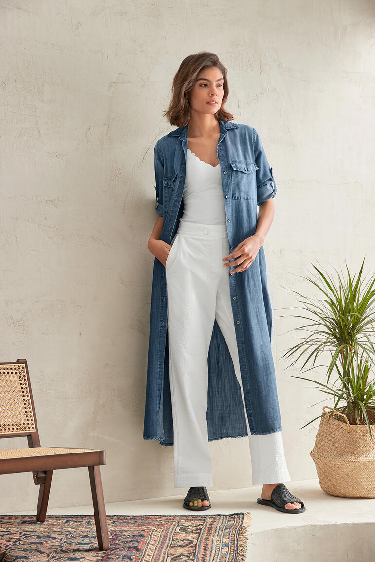 Vrouw in een jeansblousen jurk staat bij een muur met plant.