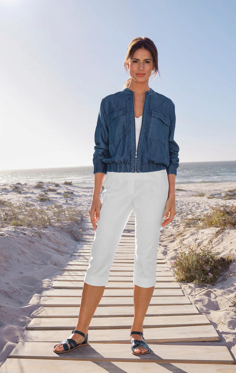 Vrouw in light-denim blouson en witte capri-broek staat op het strand.