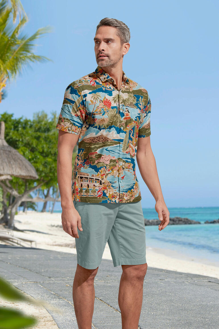 Man in een kleurig Aloha-shirt staat op het strand, omringd door palmen.