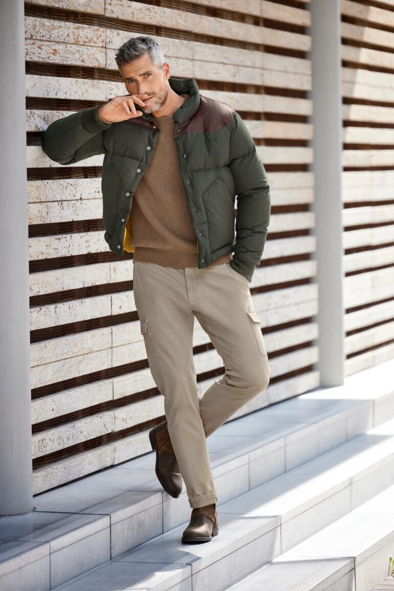 Schott NYC rangerjack Man in kamelenhaar pullover en jas, zittend op stenen.