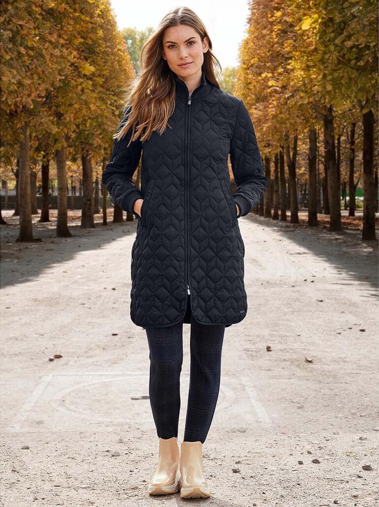 Vrouw in een zwarte gewatteerde jas staat voor de achtergrond van een park.