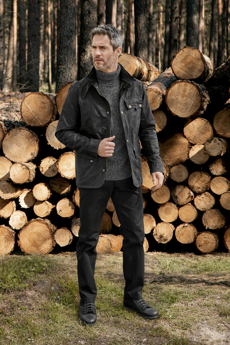 Man in waxed cotton jas staat in een bos voor stammen.