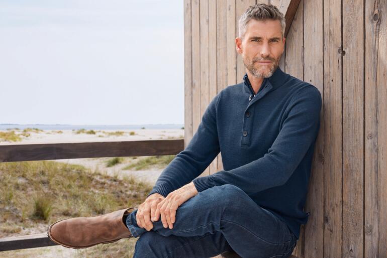 Man in een marineblauwe trui met korte kraag op een terras.