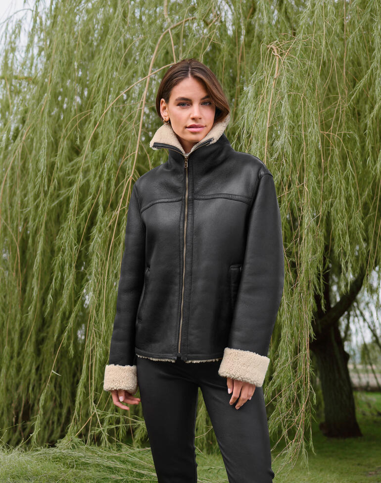 Vrouw draagt een zwart curly lamsvacht pilotenjack voor een groene wilgenboom.