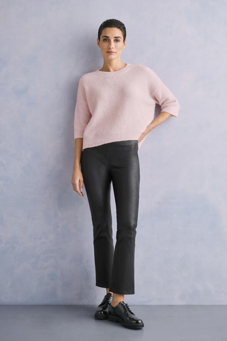 lichtpink Vrouw in roze trui en zwarte kick-flare leren broek voor een grijze achtergrond.