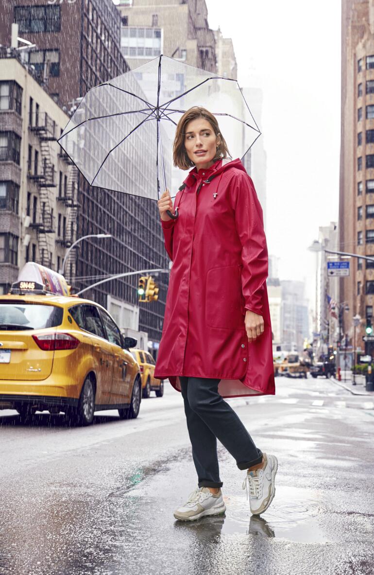 Ilse Jacobsen Red Raincoat Vrouw in een rode regenjas houdt een doorzichtige paraplu in een stedelijke omgeving.