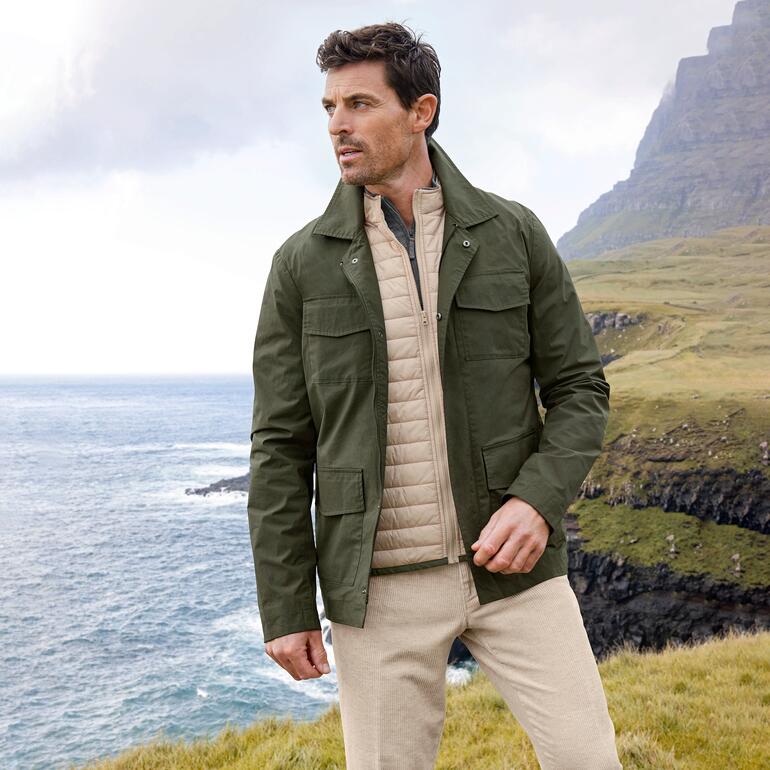 Weerbestendig fieldjacket Man in weerbestendige fieldjacket staat aan de kust met een groene berg op de achtergrond.