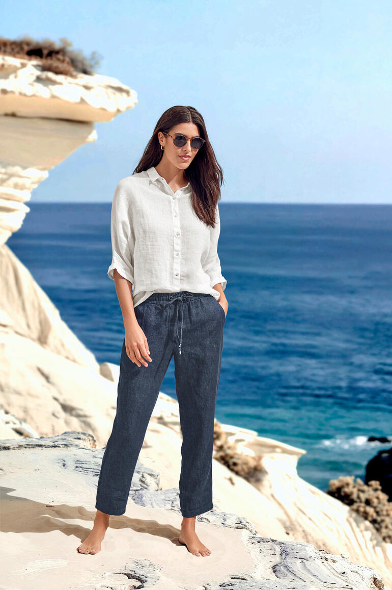 Vrouw in luchtige blouse en broek staat aan de zee.