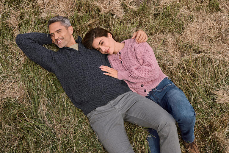 heren, navy en dames, lichtroze Man en vrouw liggen op een grasveld en dragen gebreide Aran-cardigans.