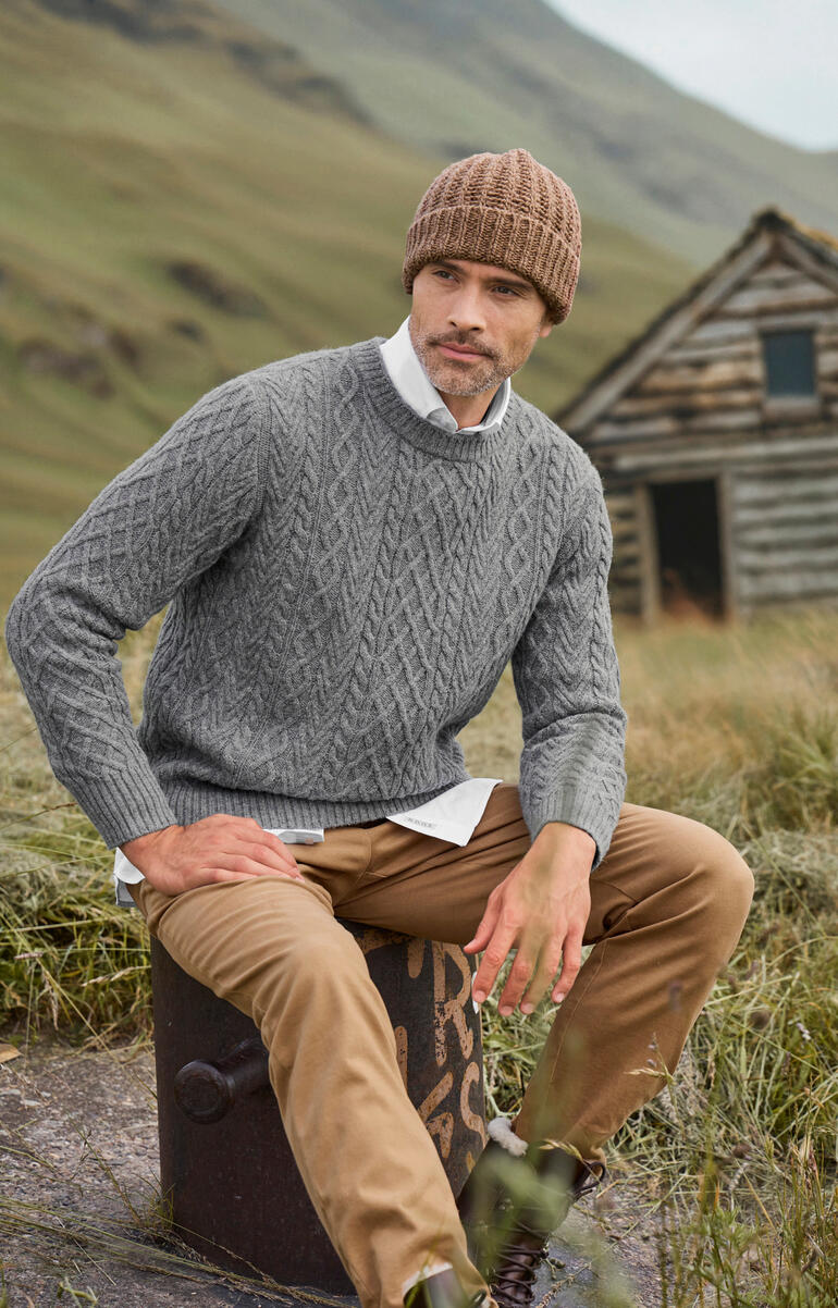 Duneen Ireland Aran-trui Man zit op een steen, draagt een grijze Aran-trui en een muts.