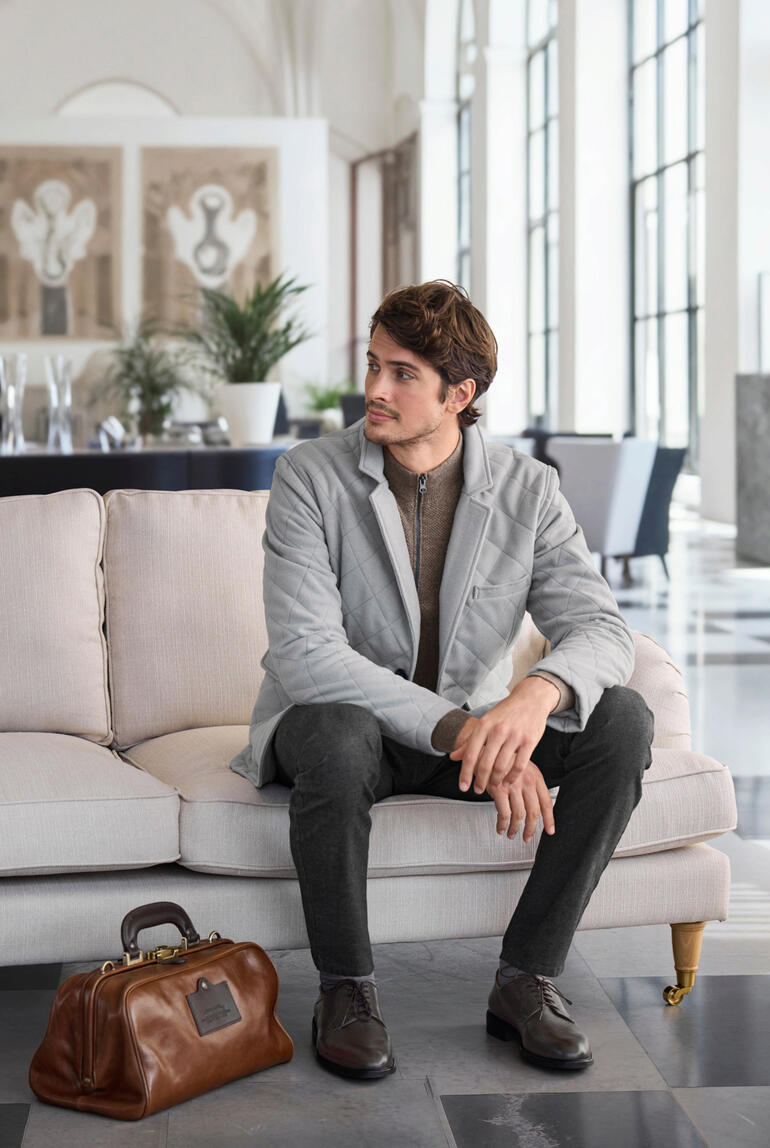 Man met grijze kasjmier gewatteerde jas zit op een bank, elegante tas naast hem.