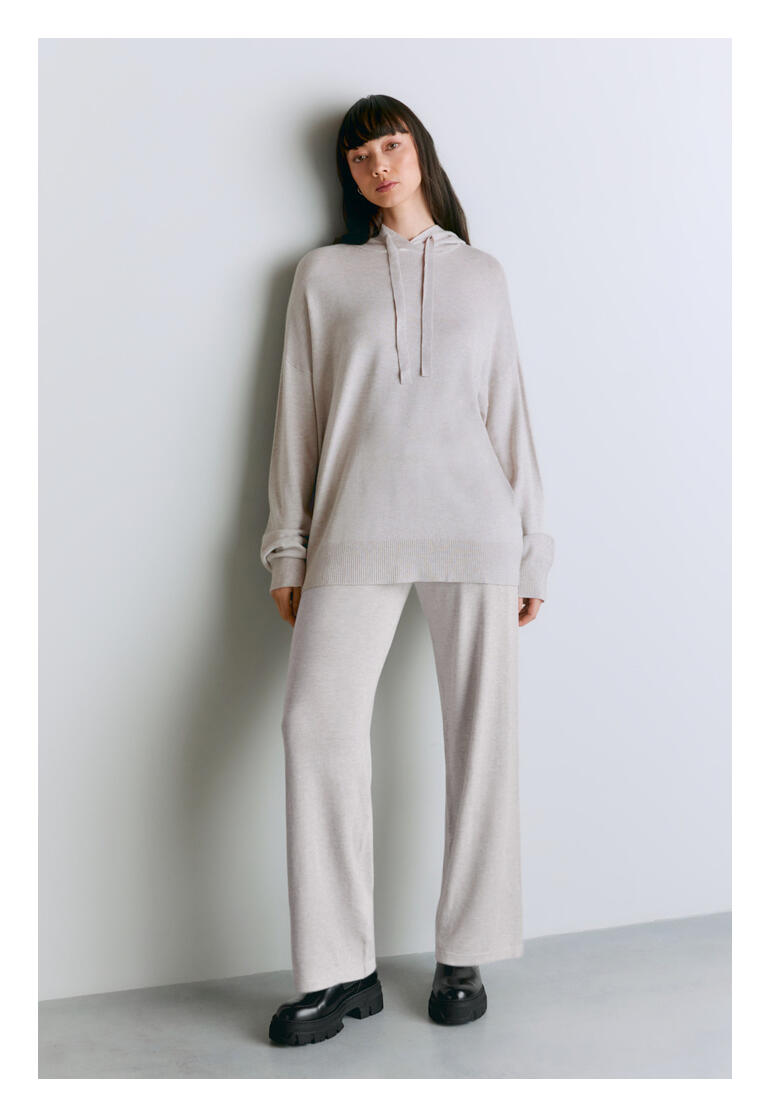Vrouw in een lichtgrijze lyocell/kasjmier hoodie, staande voor een muur.
