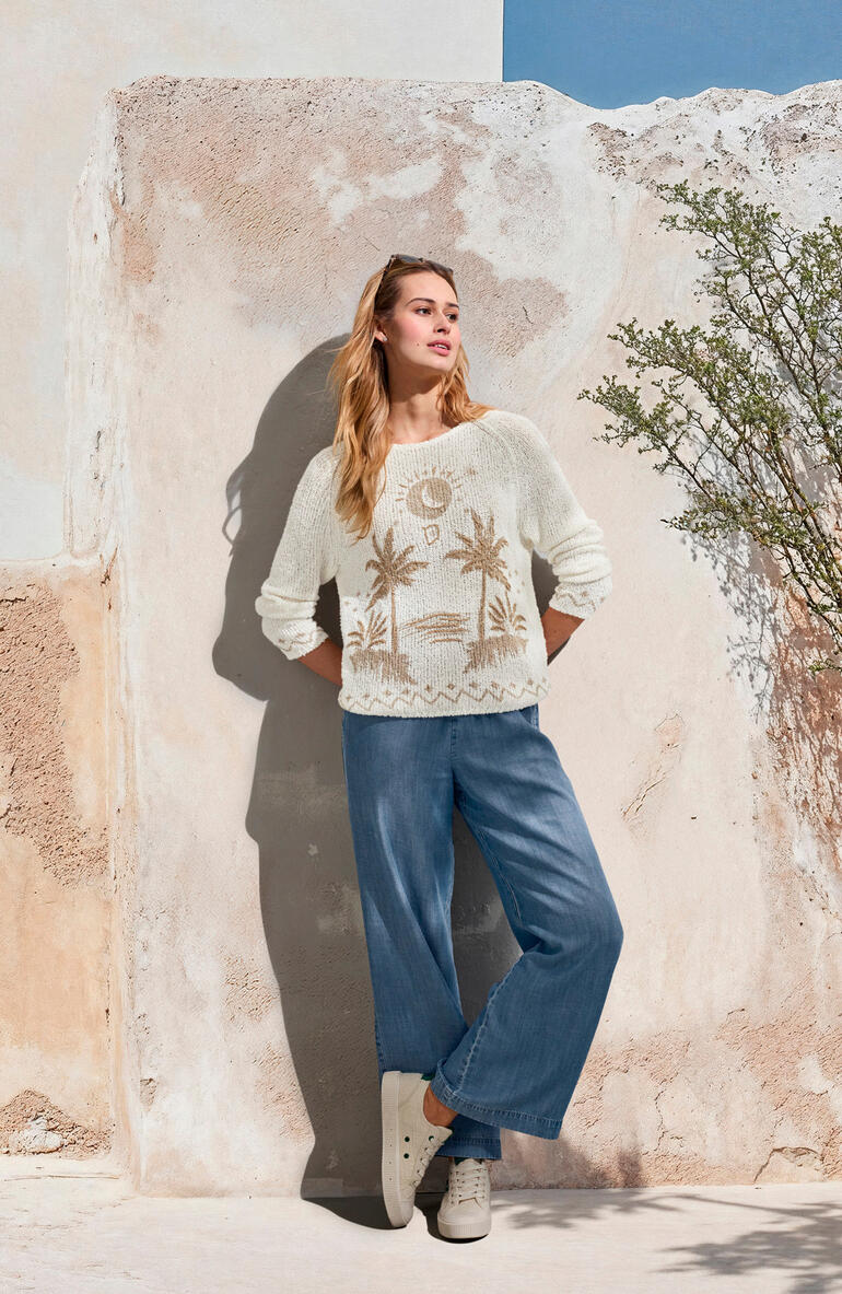 Vrouw in crème kleurige trui en lichtblauwe wijde jeans, voor een muur.