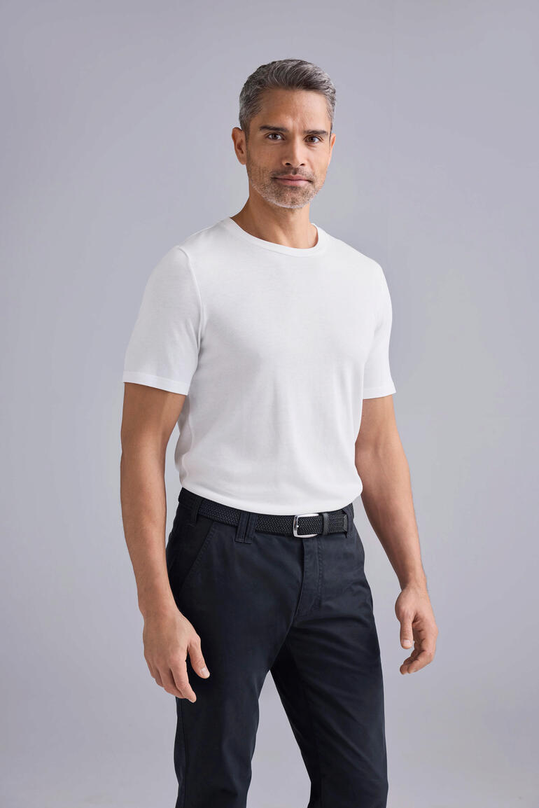 Man in een wit T-shirt en zwarte broek, staat voor een grijze achtergrond.