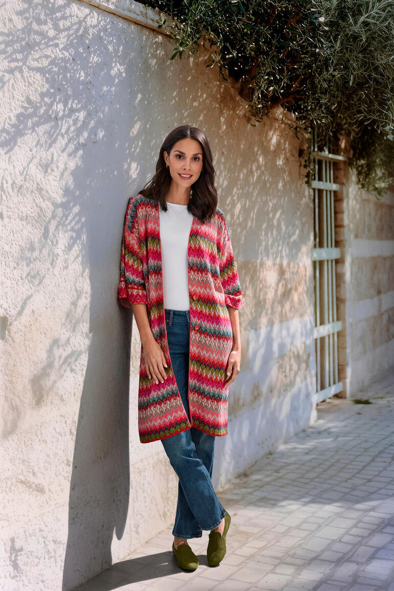IVKO lange jacquard-cardigan Vrouw in een kleurrijke gebreide lange cardigan met gekleurde ruitpatronen, staat tegen een muur.