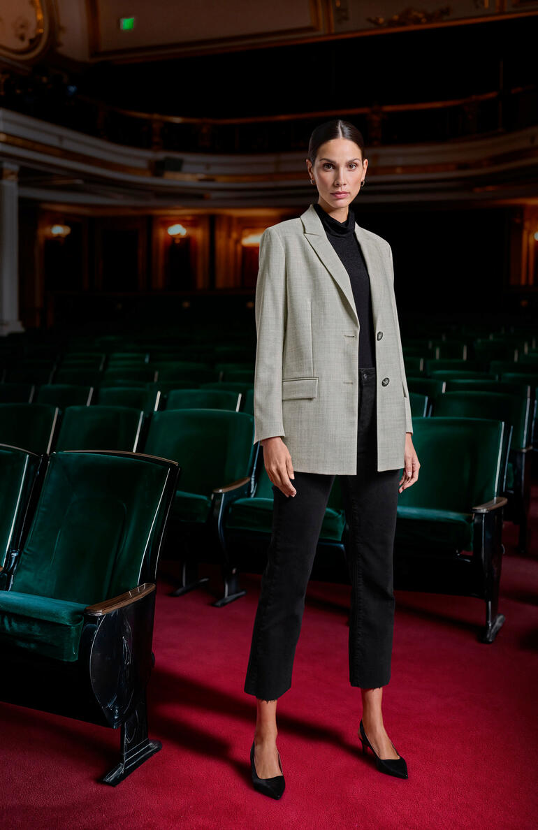 Vrouw in een geribbeld zwart topje en grijze blazer staat in een theater.