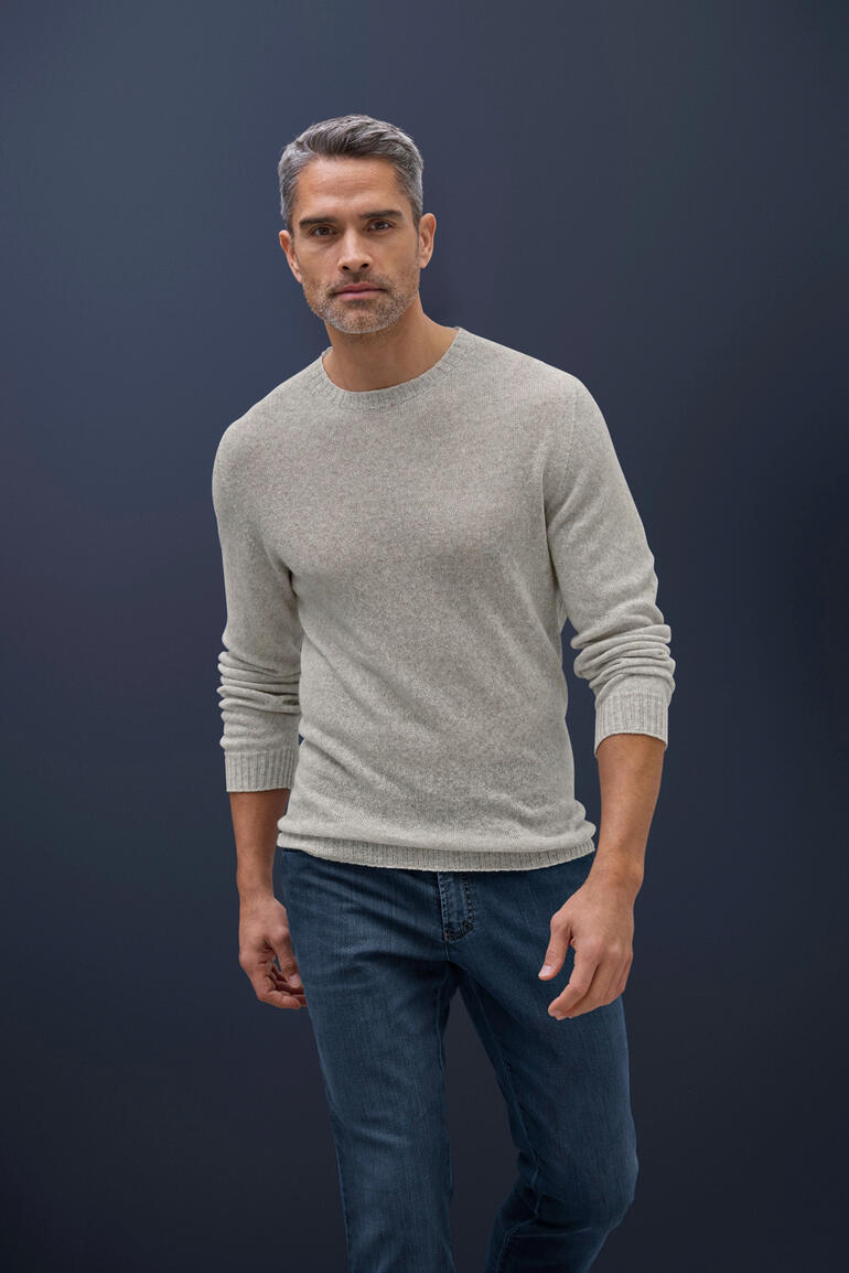 taupe Man draagt een lichtgrijze kasjmier-linnen trui en jeans op een donkere achtergrond.