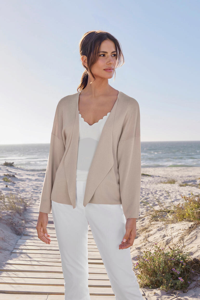 Vrouw draagt een waterfall cardigan op het strand, zon schijnt.