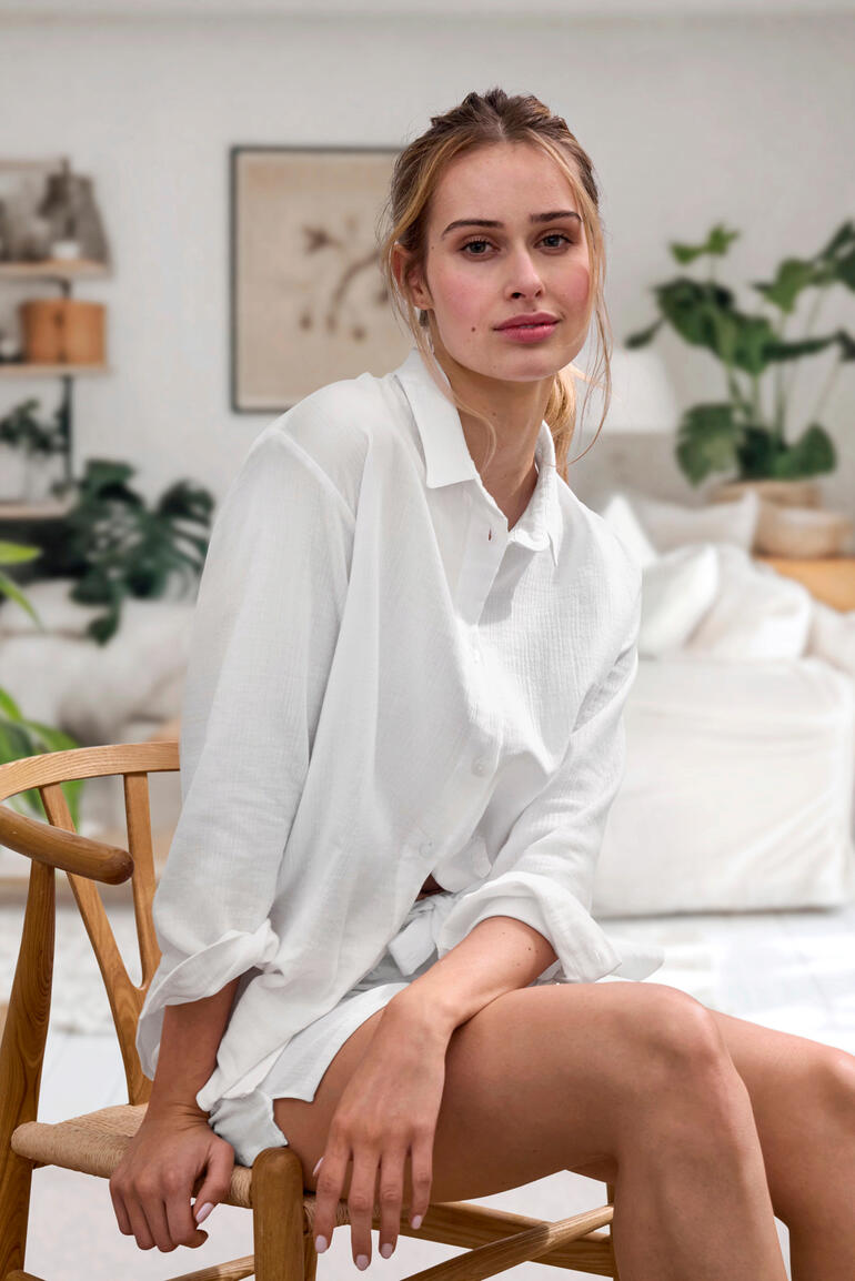 blouse en short Vrouw zit op een stoel, draagt een witte Novila Musselin blouse, met natuurlijke planten op de achtergrond.
