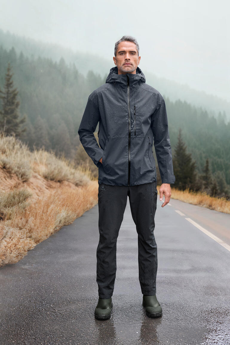 Man in grijze Hardshell-jack staat op een natte weg met bergen op de achtergrond.