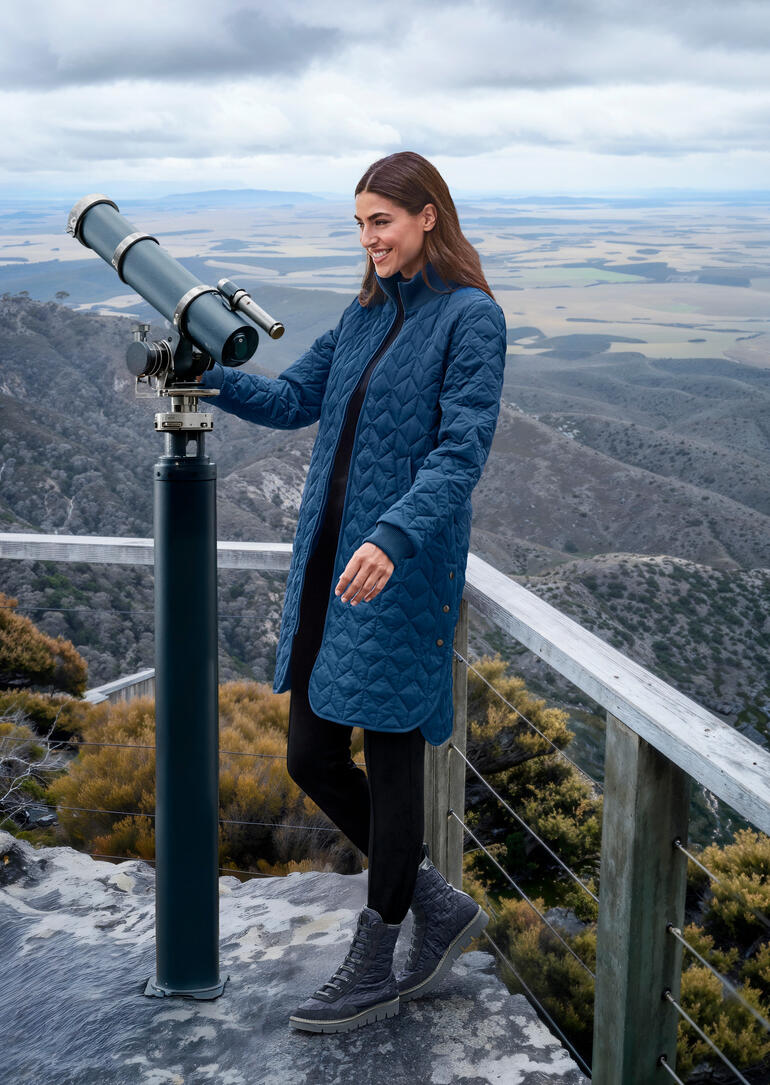 Vrouw in koninklijk blauwe gewatteerde jas, staand bij een telescoop.
