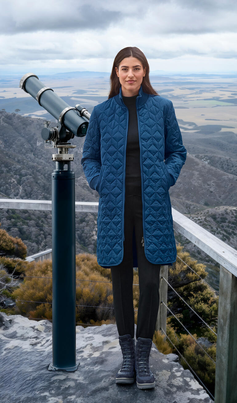Vrouw in een koningsblauwe gewatteerde jas op een uitkijkpunt met telescoop.