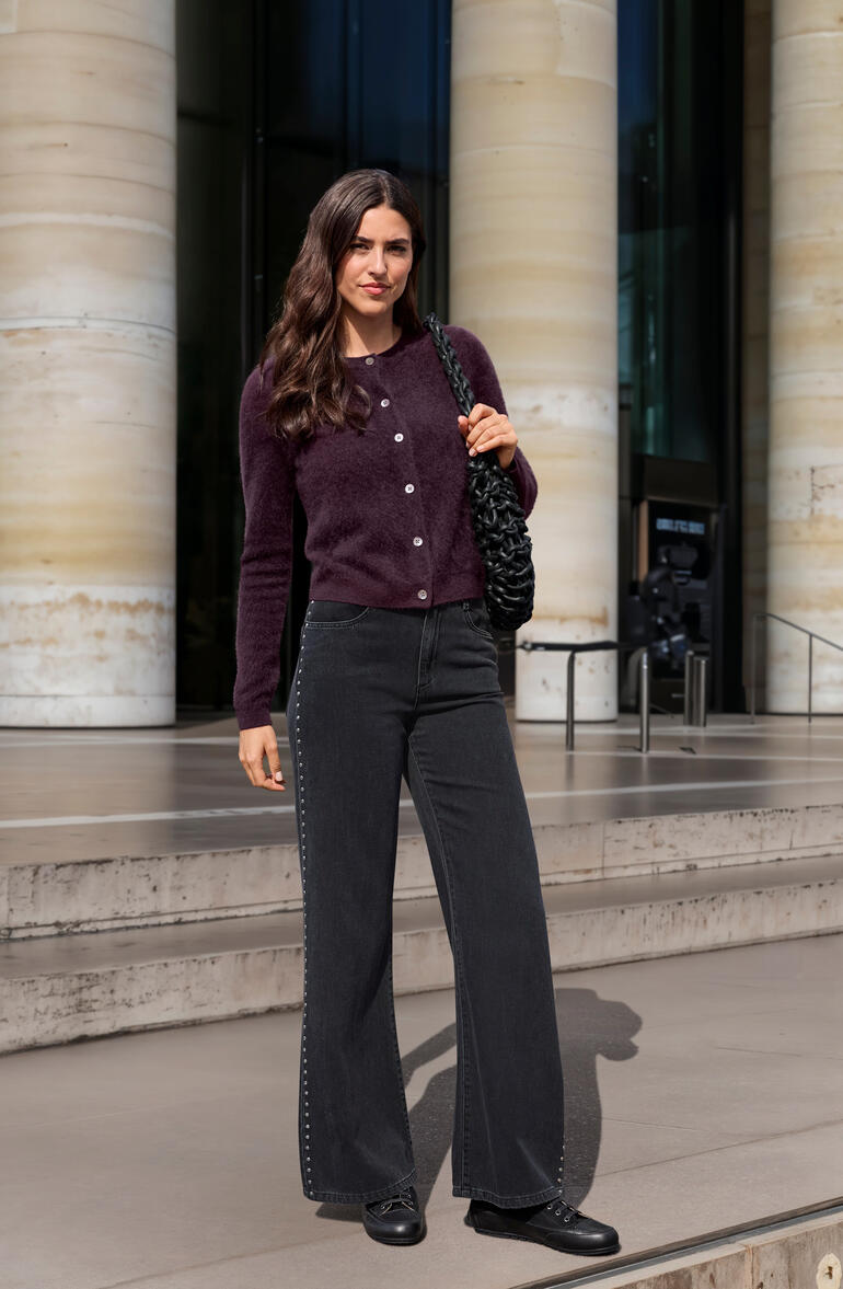 Vrouw in bordeauxkleurige cardigan en zwarte broek voor een modern gebouw.