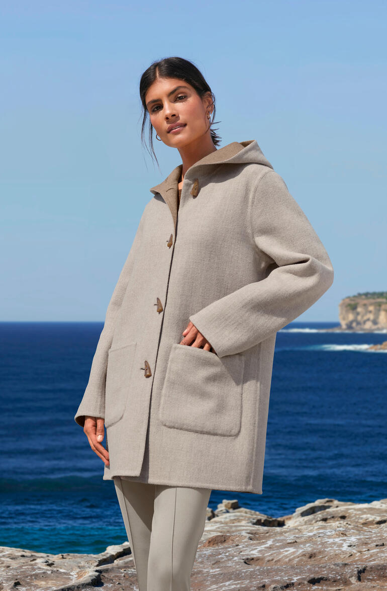Alpaca double face dufflecoat in beige, gedragen door een vrouw met de zee op de achtergrond.