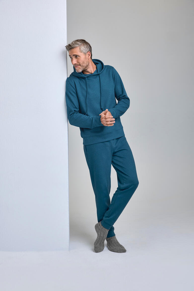 Man in tealkleurig cashmere-katoenen loungewear-pak leunt tegen de muur.