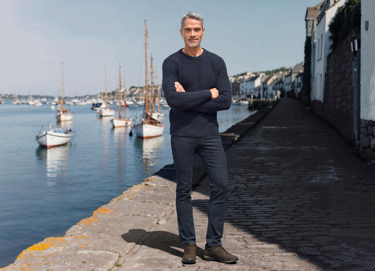 Man in een donkerblauwe ribbeltrui, staat aan de haven met boten op de achtergrond.
