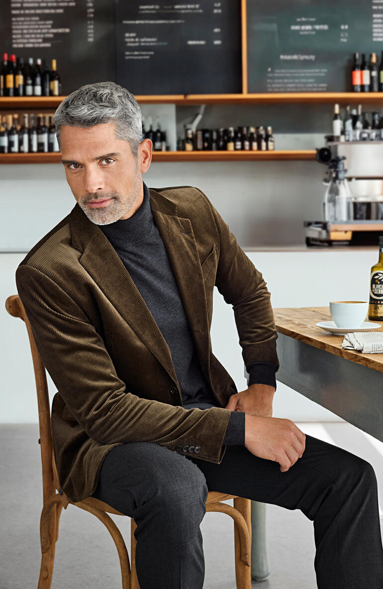 Man in een bruine corduroy blazer zit in een café met een elegante uitstraling.